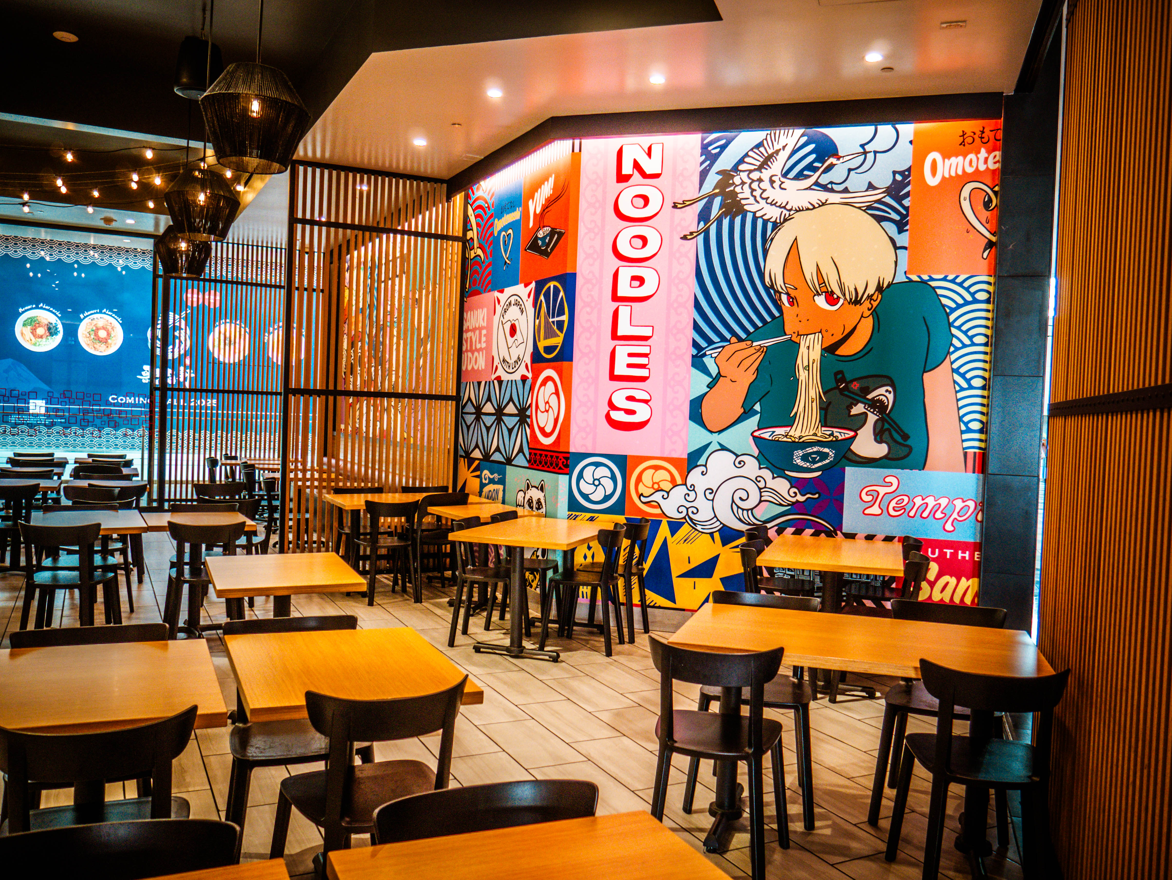 Dining area featuring vibrant anime character mural with blonde character eating noodles, wooden tables, and prominent 'NOODLES' signage