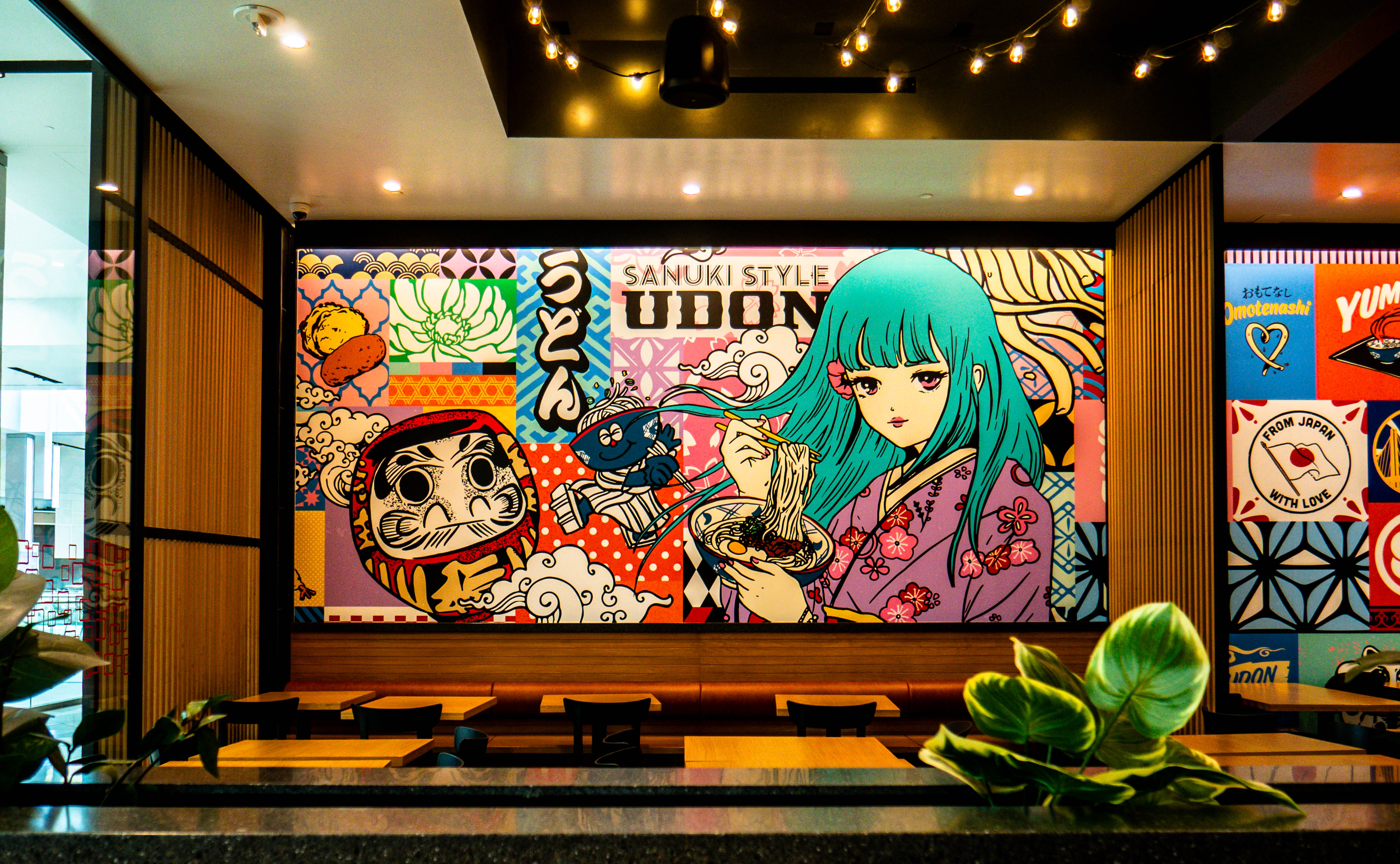 Dining area with large anime character mural featuring teal-haired girl and Daruma doll, wooden banquette seating, and ambient string lighting