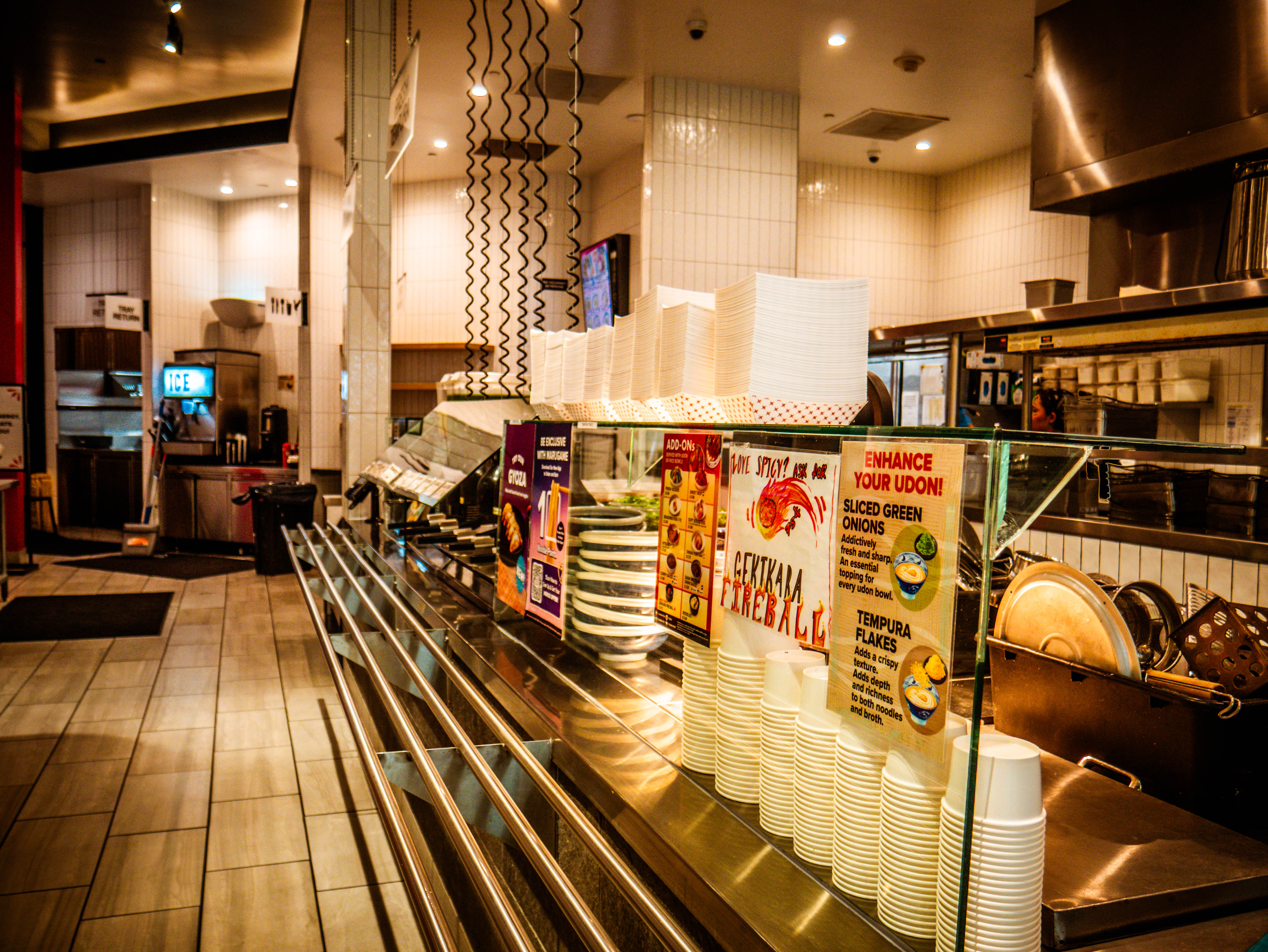 Self-service condiment line with 'ENHANCE YOUR UDON!' signage, cups, and customer enhancement stations