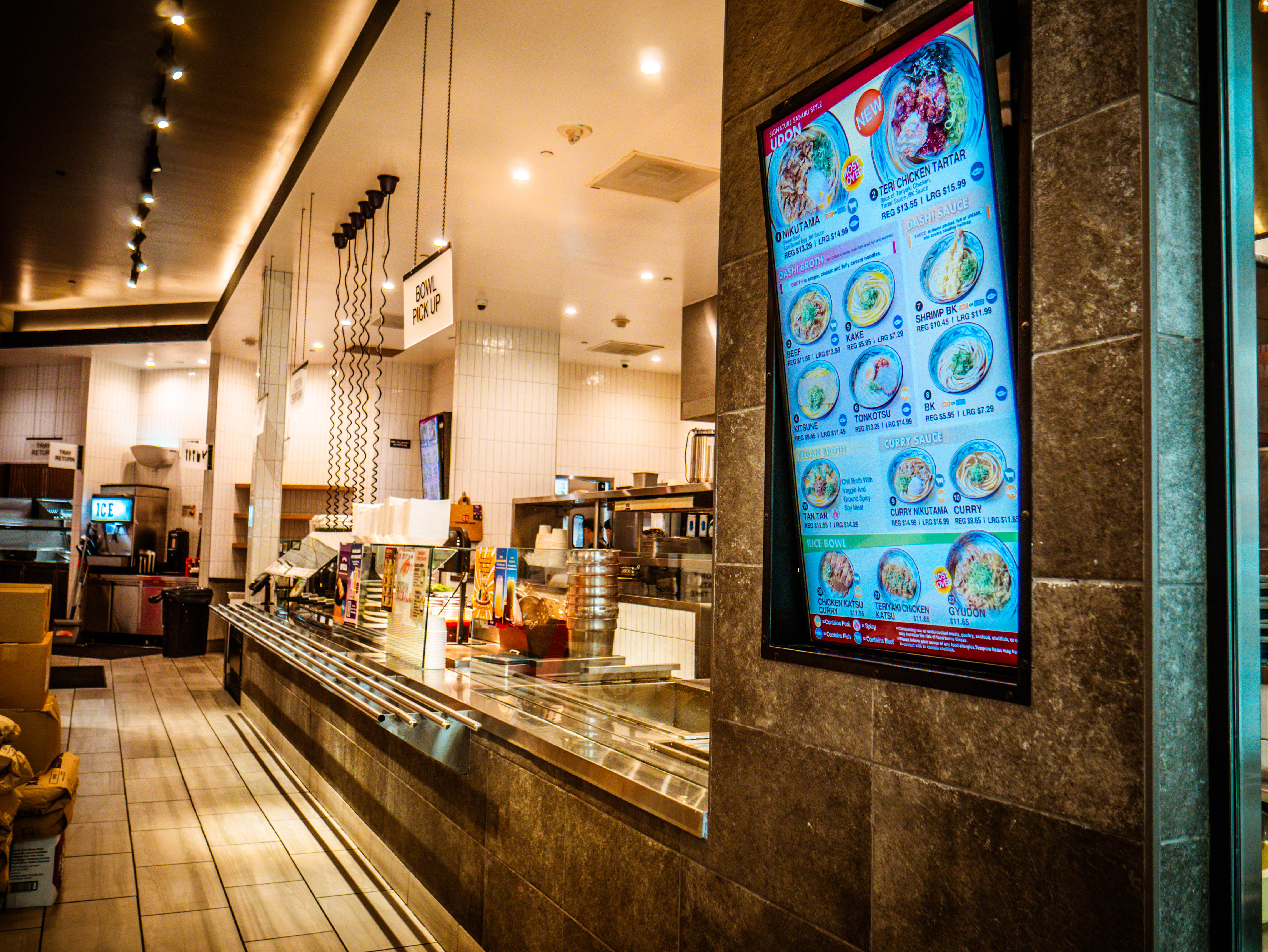 Large digital menu board displaying colorful udon dishes with view of open kitchen and professional service counter