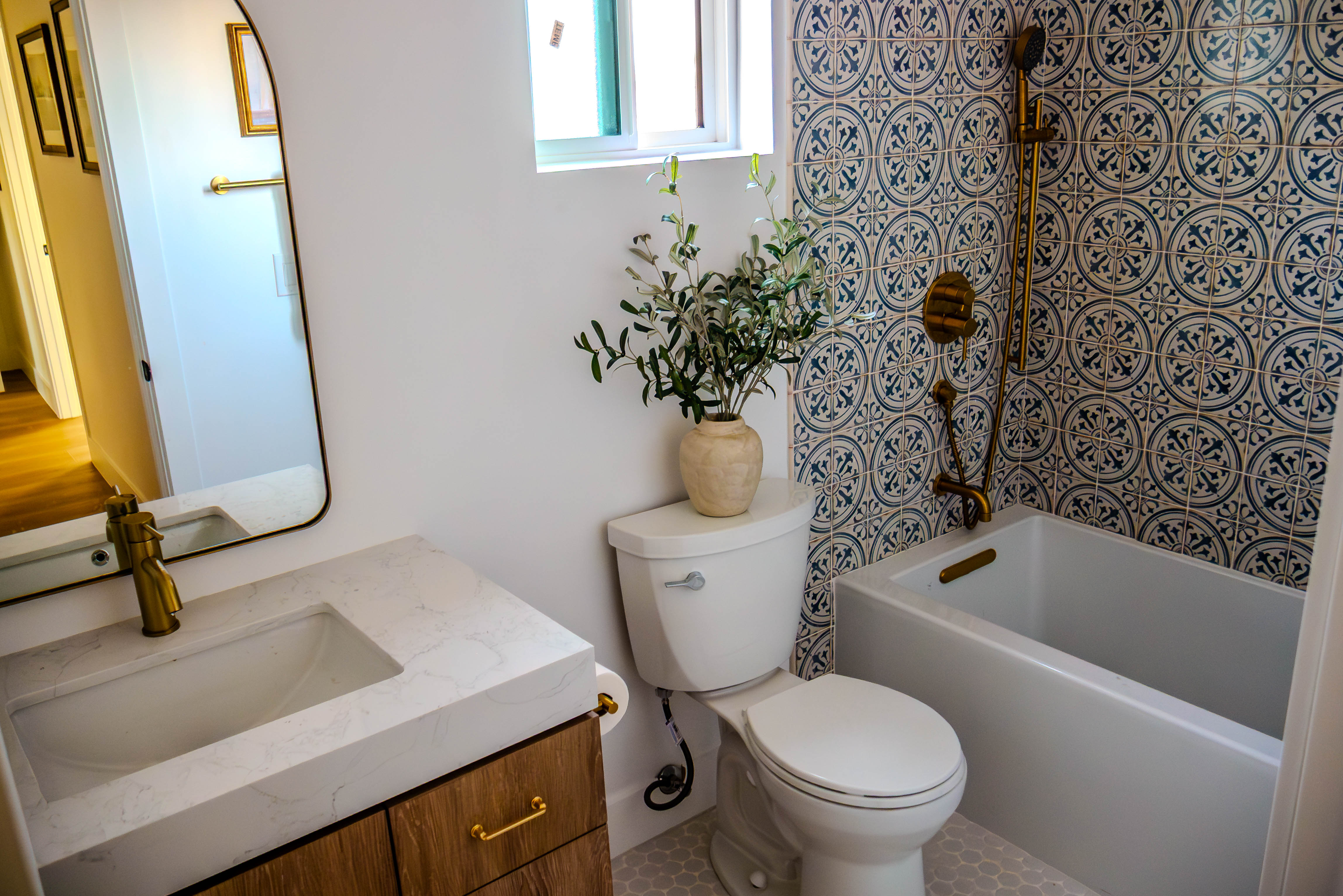 Modern bathroom with striking blue and white geometric patterned tile wall, white vanity with natural wood doors, brass fixtures, and hexagonal floor tile