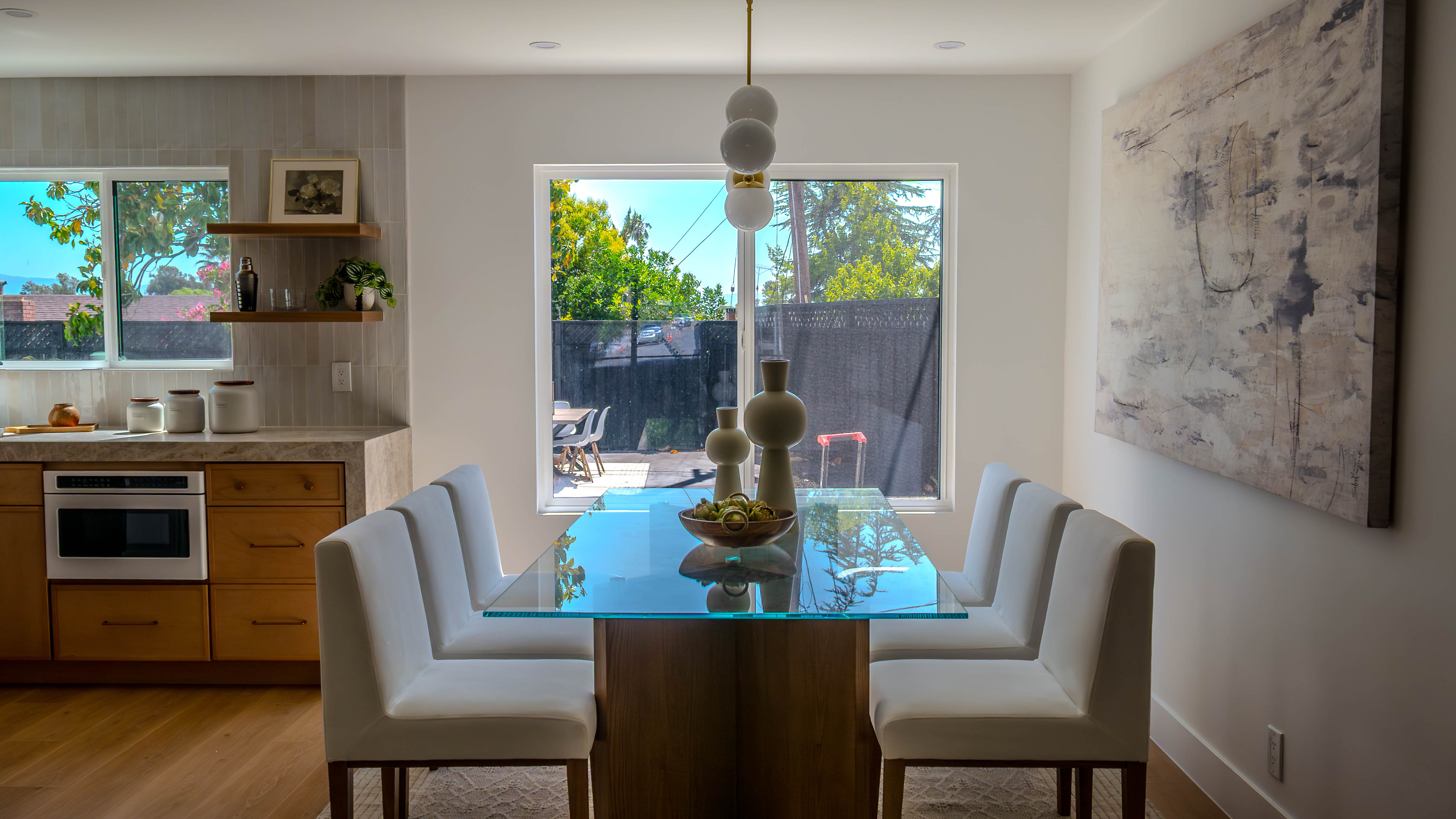 Dining area with glass-top table, white upholstered chairs, contemporary artwork, and modern pendant lighting