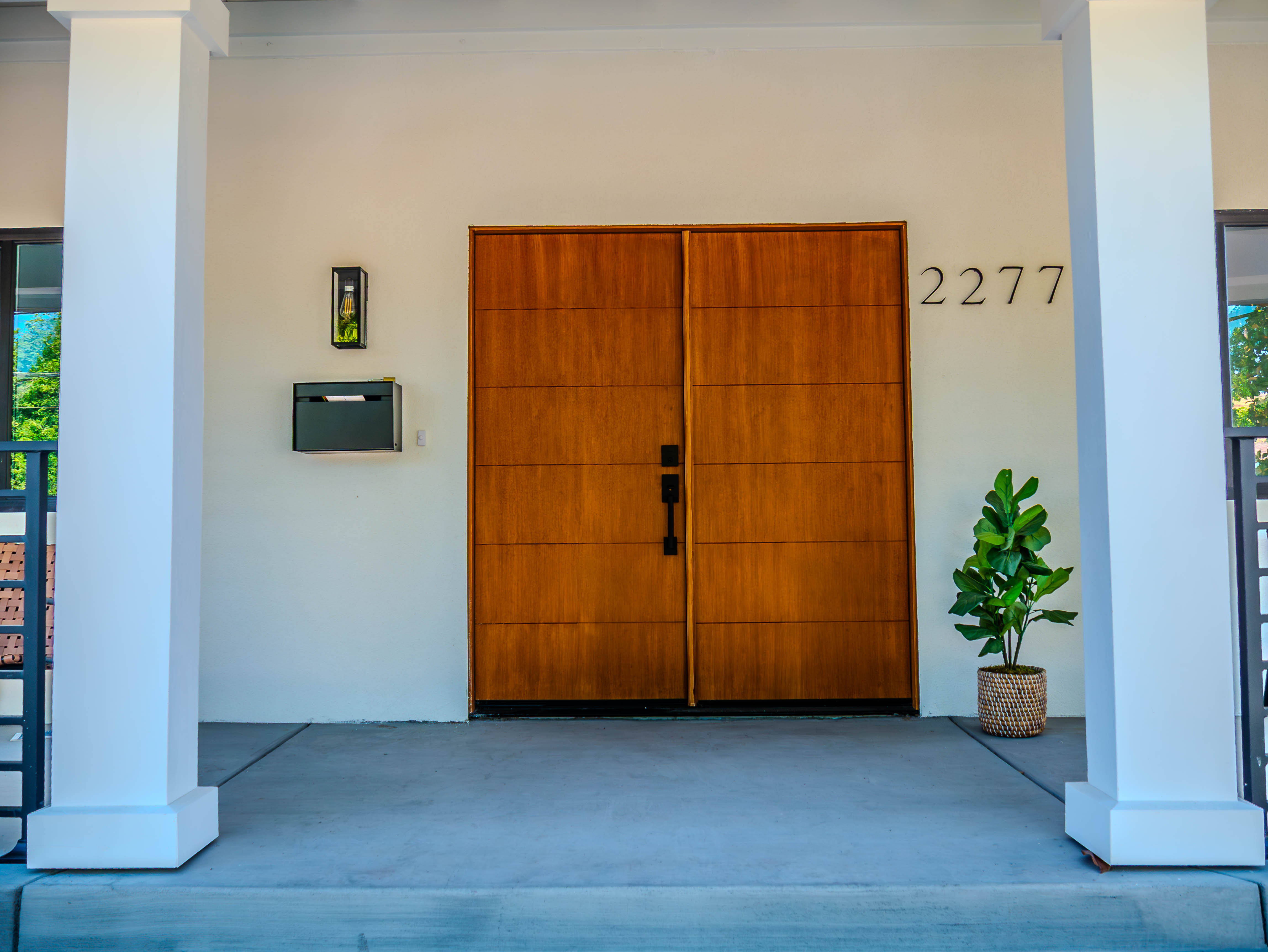 Close-up of natural wood double doors with house number 2277, modern mailbox, and decorative plant