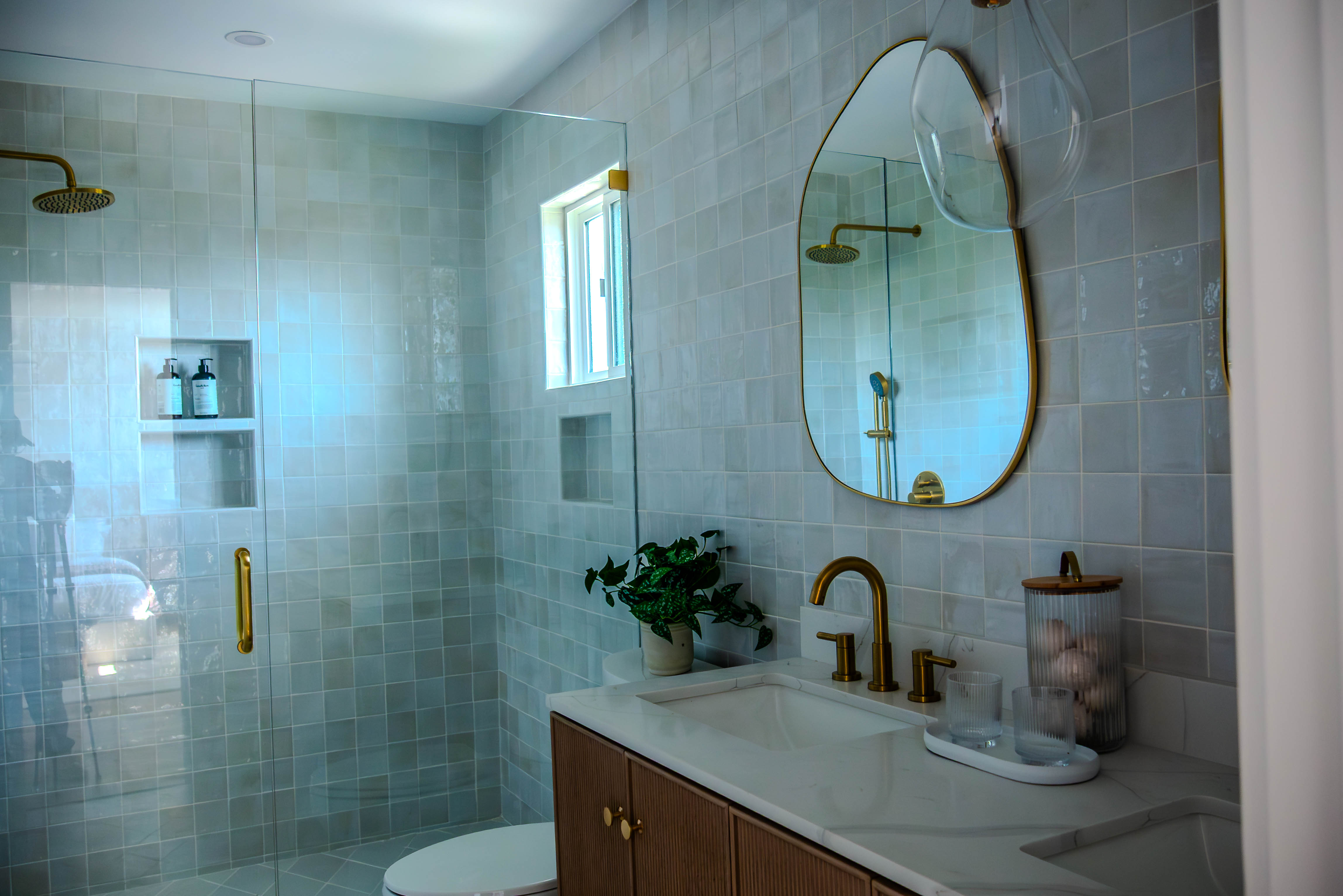 Spa-like bathroom with sage green tile, brass fixtures, rainfall shower, and oval mirror