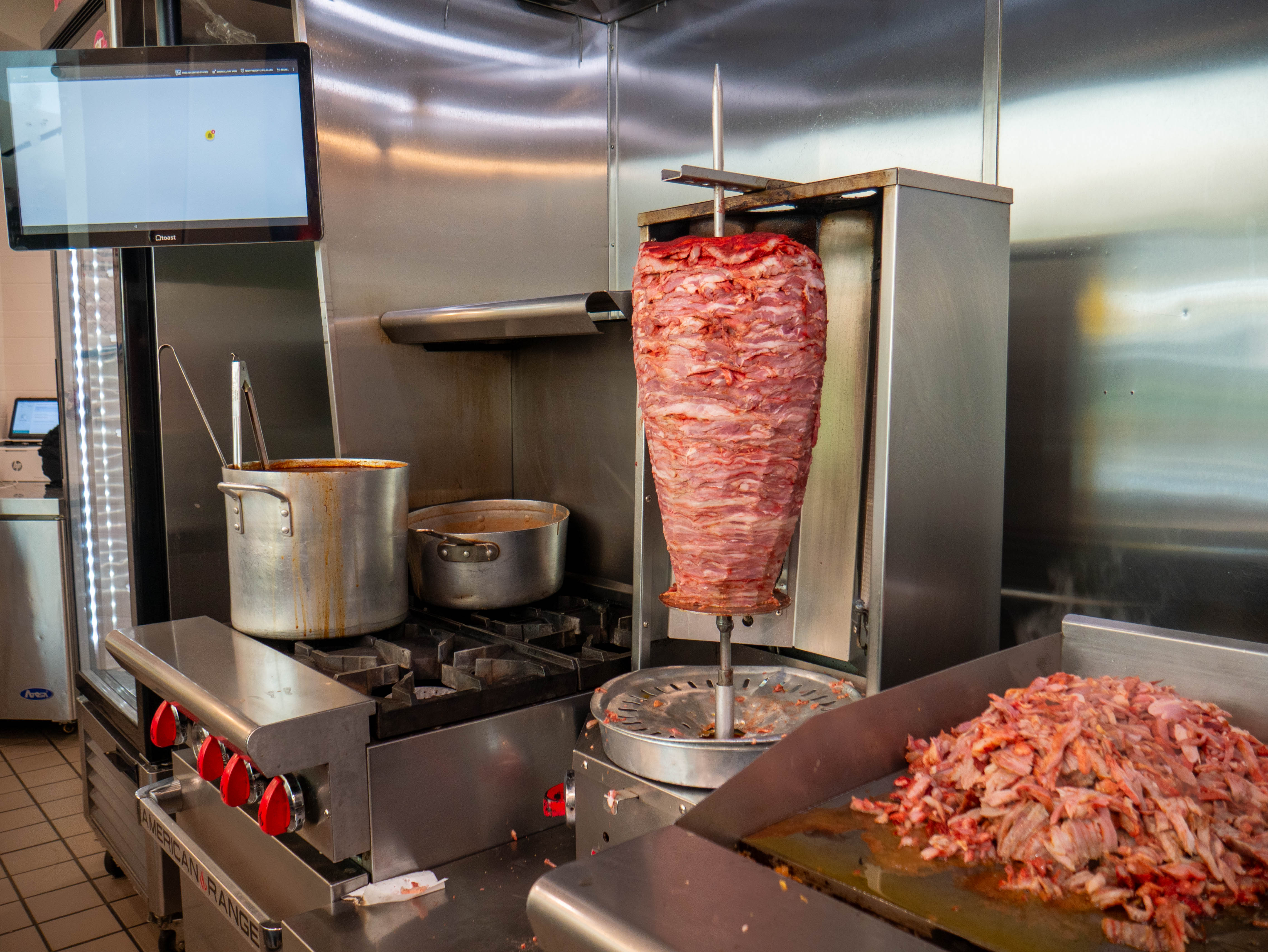 Professional kitchen featuring traditional al pastor trompo with vertical meat spit, commercial stove, and authentic Mexican cooking setup