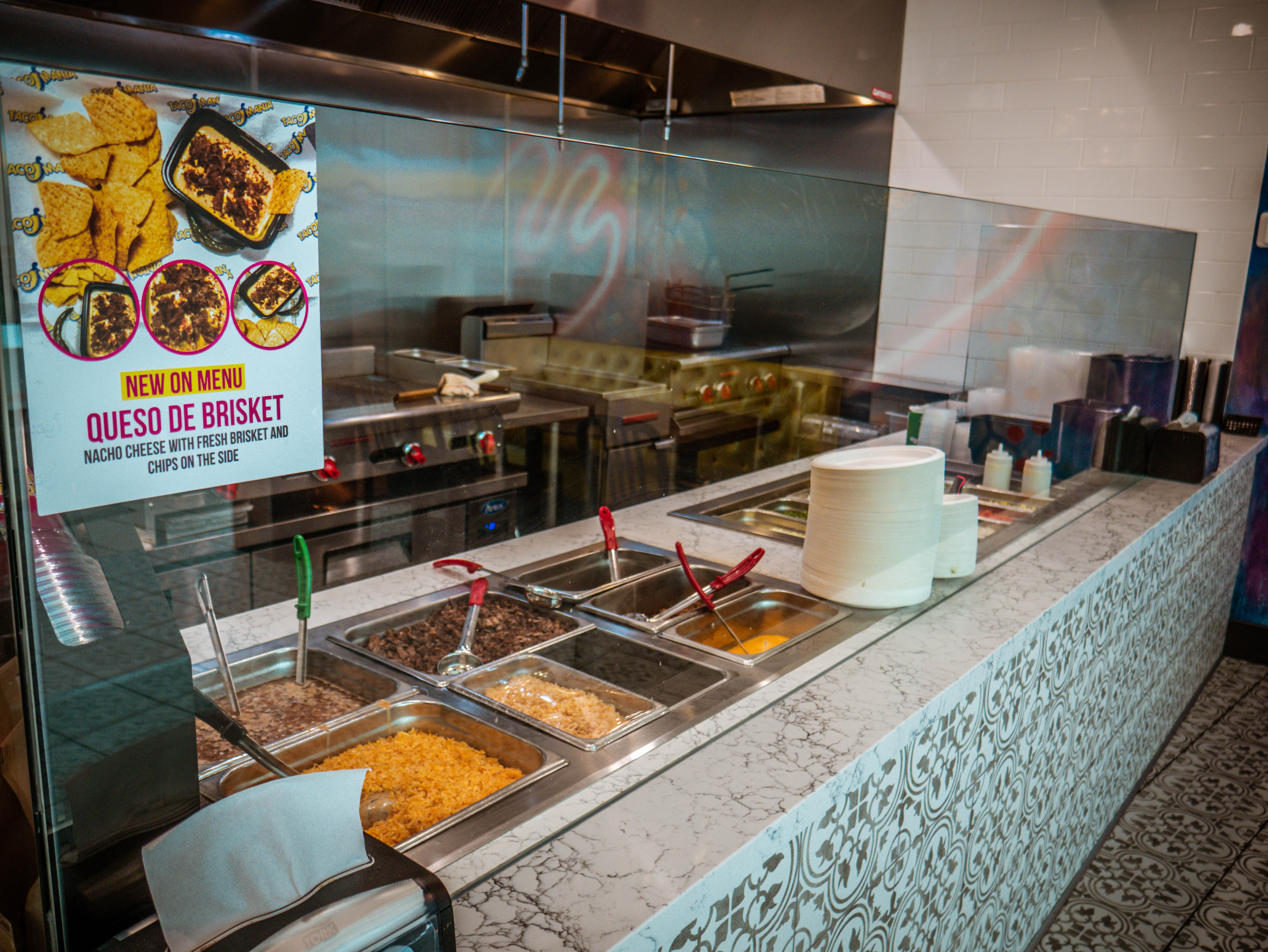Professional food service line with colorful ingredient stations, stainless steel containers, and promotional signage for Queso de Brisket