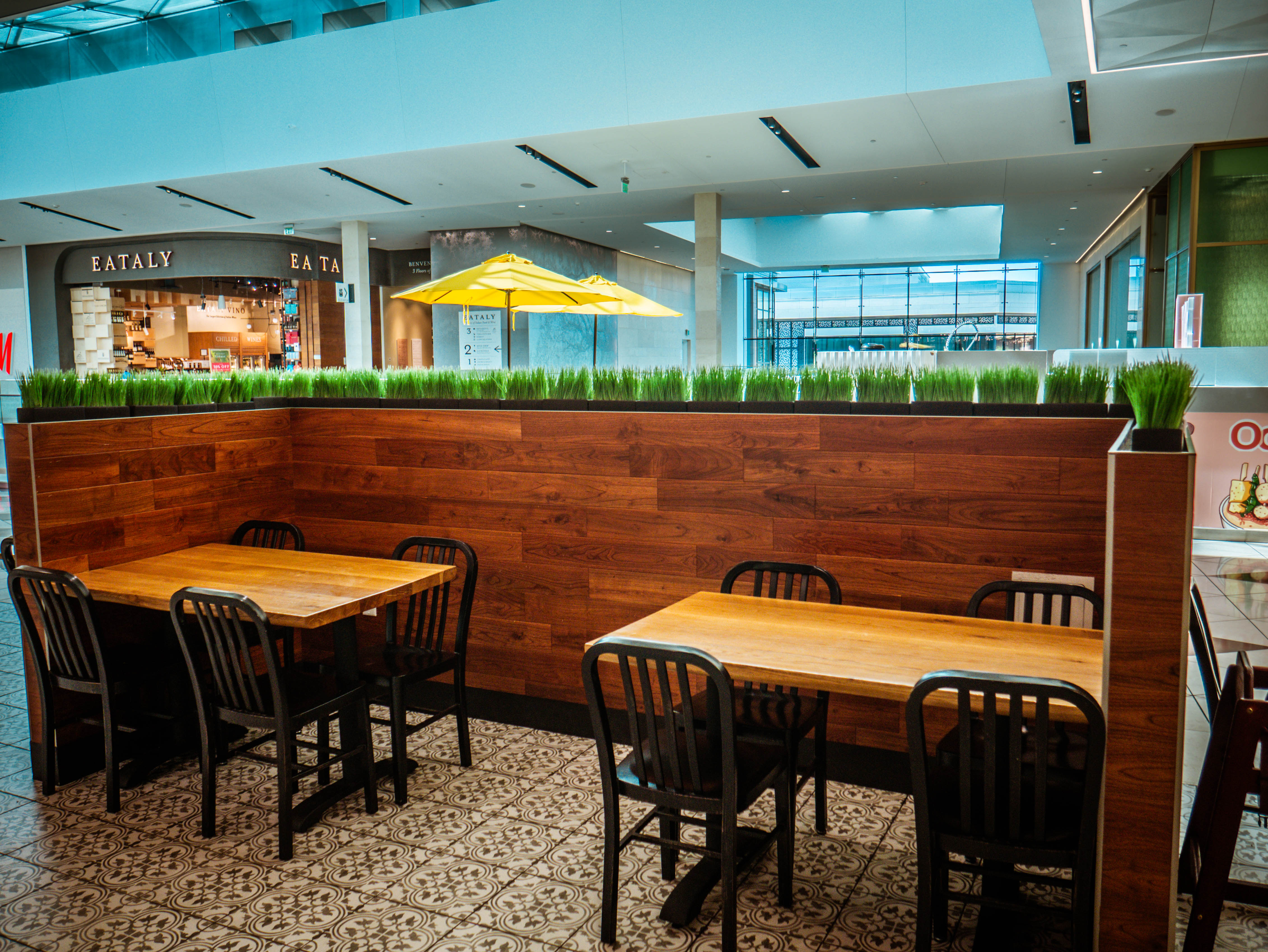 Mall-facing seating with wooden tables, decorative grass planters, and seamless integration with Westfield Valley Fair corridor