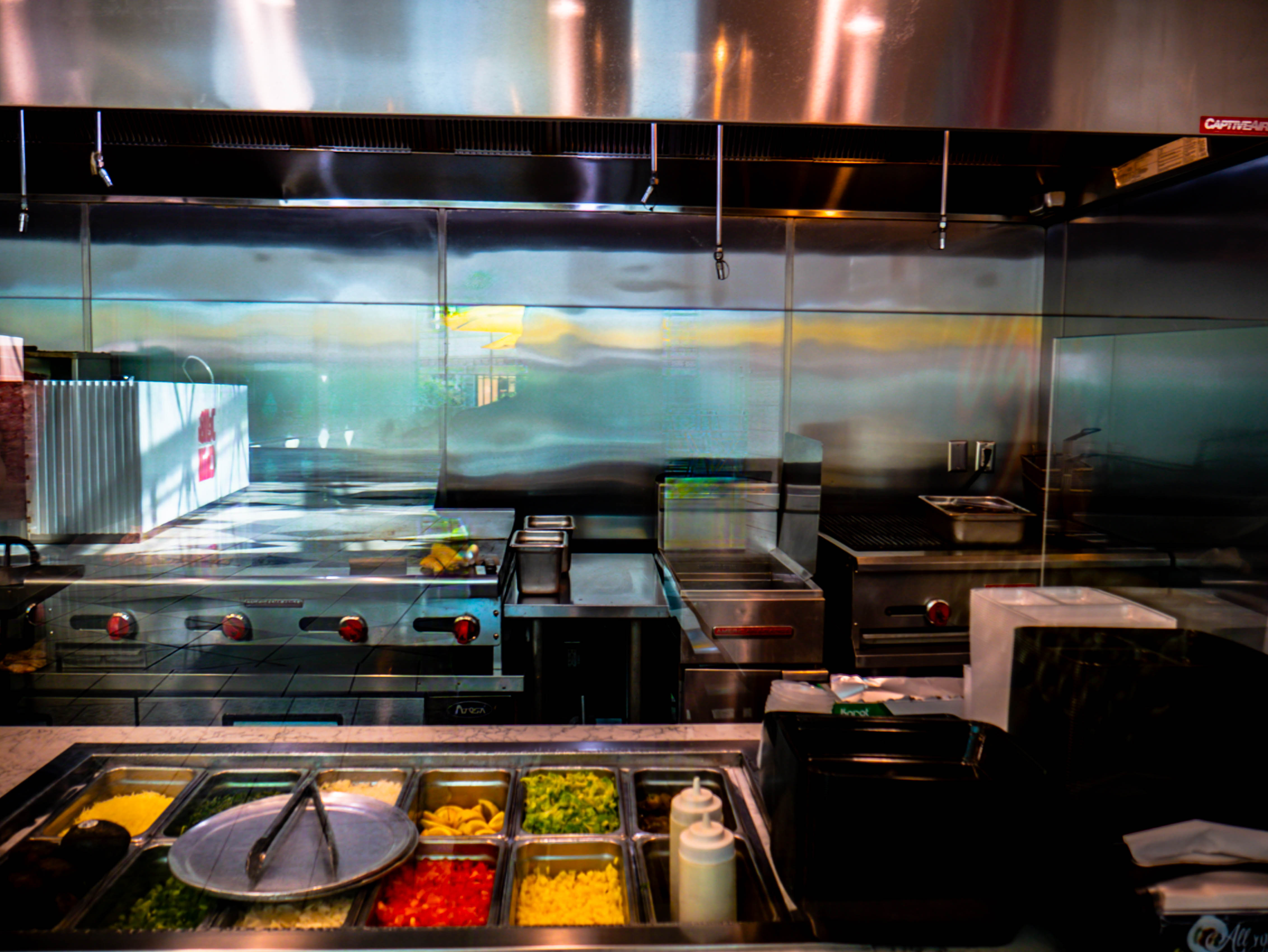 Professional commercial kitchen with stainless steel equipment, ingredient prep stations with colorful toppings, and grills