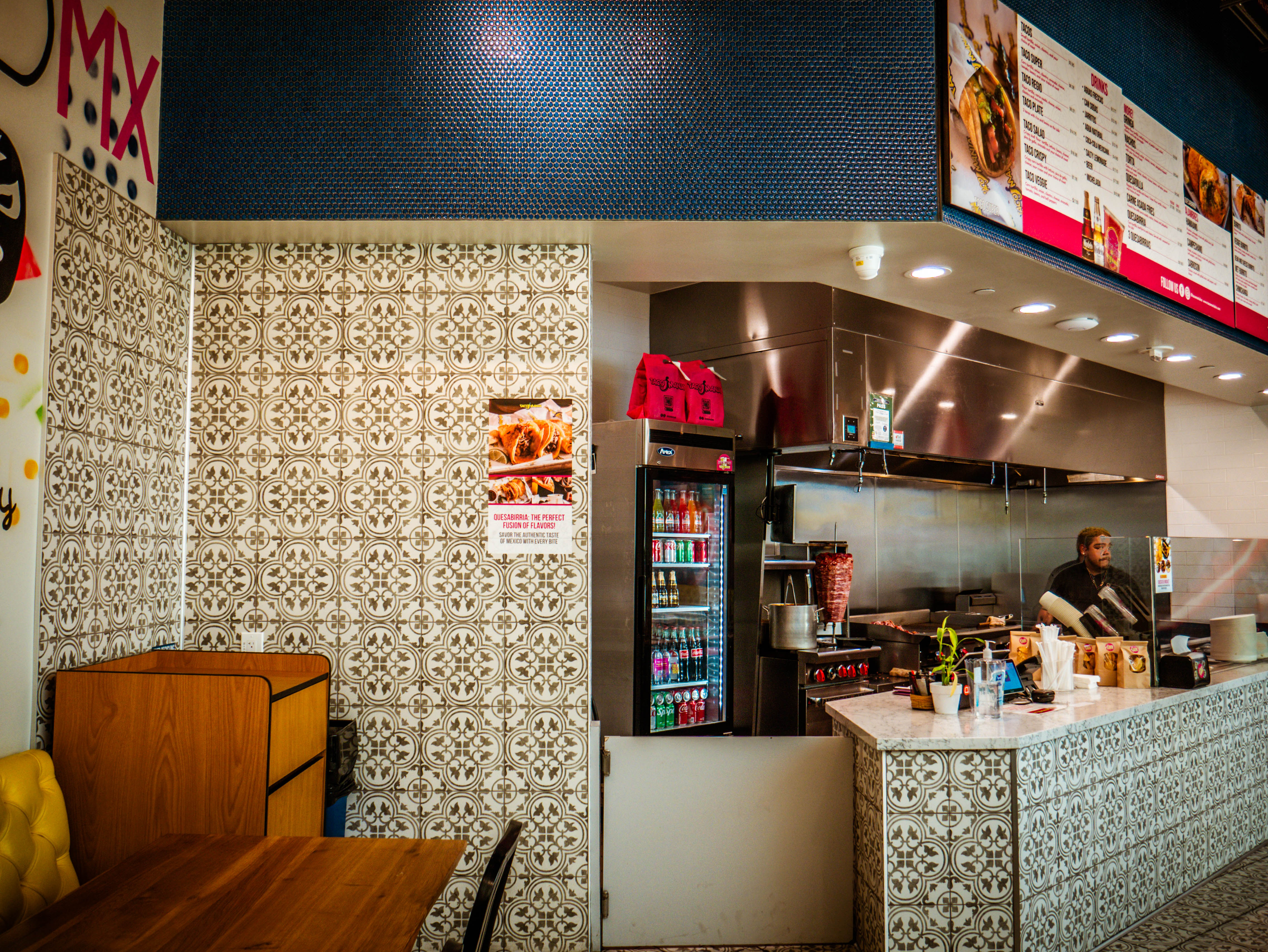Interior service counter with decorative tile work, open kitchen area, beverage cooler, and staff member working in fast-casual setup