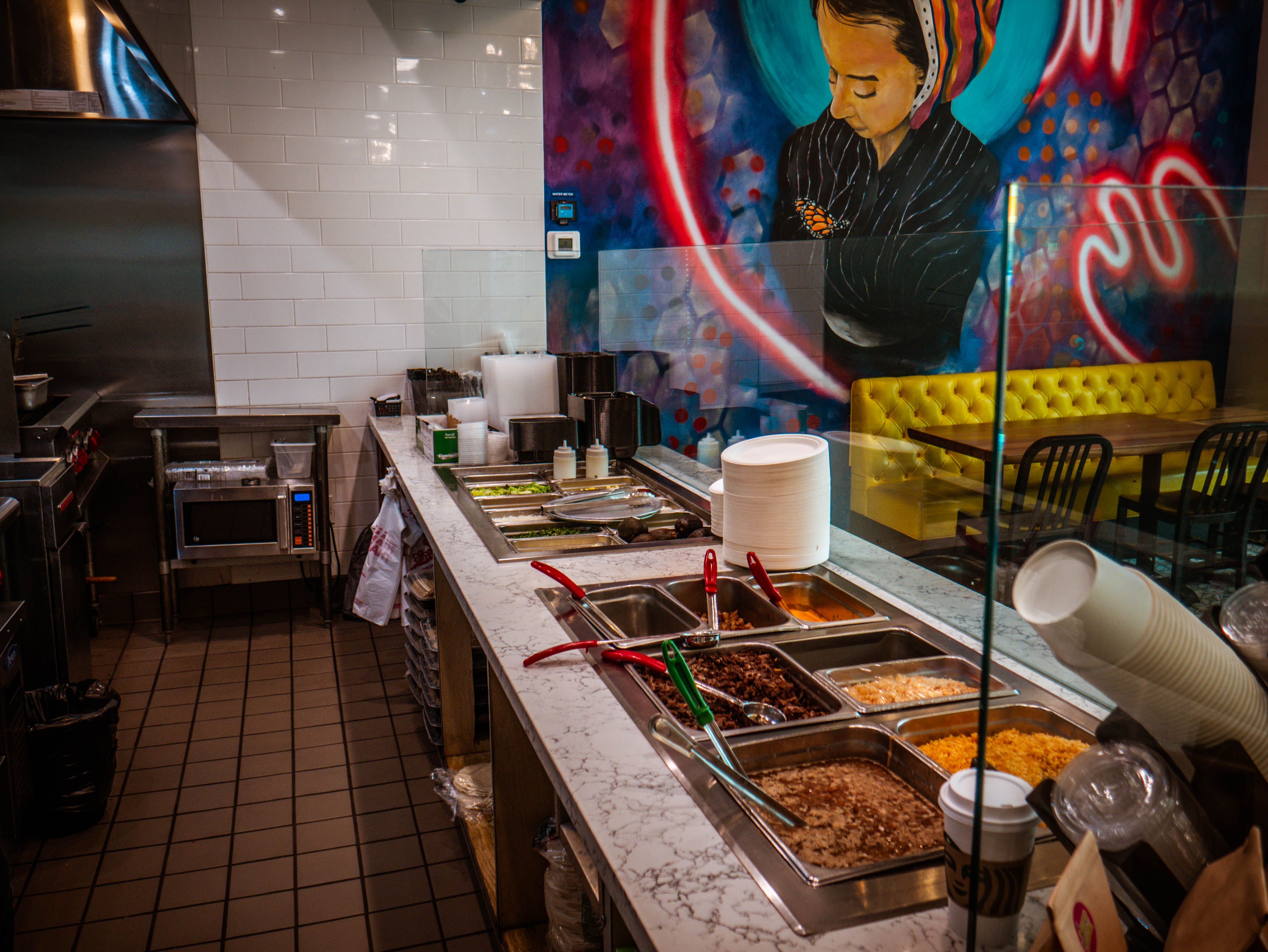 Behind-the-scenes prep area of food service line with ingredient containers, woman mural backdrop, and operational setup